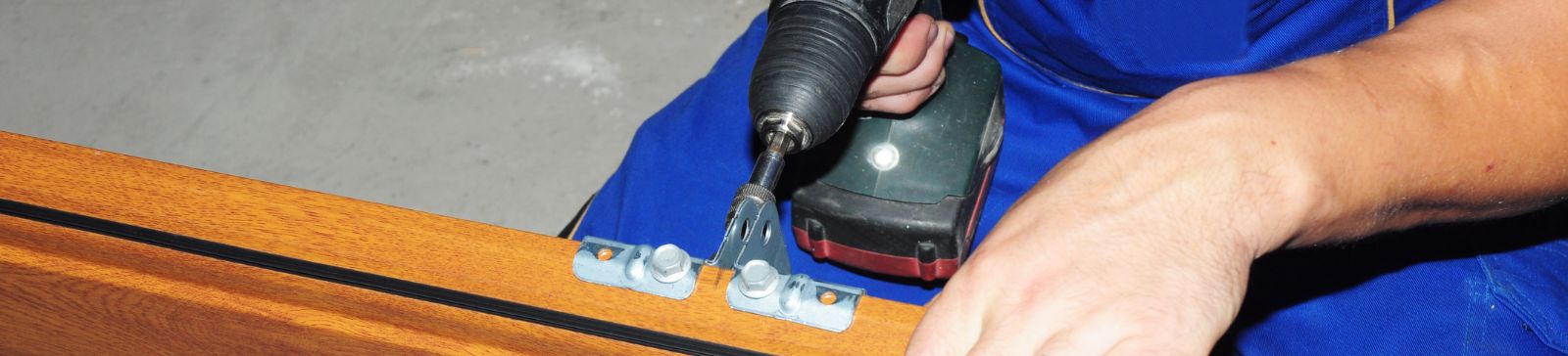 Technician in blue uniform using a power drill to repair a wooden garage door panel.